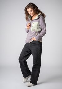 Frau mit lockigem Haar, die eine violette Fleecejacke mit einer hellgrünen Brusttasche, schwarze Jeans mit weitem Bein und weiße Turnschuhe trägt, stehend und lächelnd.