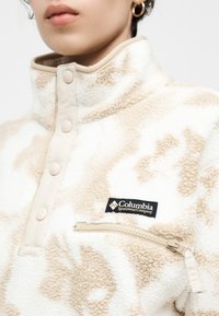 Κοντινό πλάνο ενός ατόμου που φοράει μπεζ και λευκό φλις μπουφάν Columbia με κουμπιά snap και τσέπη  στο στήθος με φερμουάρ.