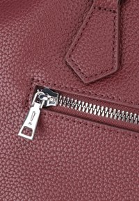 Zipper s kovinskim potegalkom na teksturirani temno rdeči usnjeni površini, ki prikazuje podrobno šivanje in nabritost usnjene zrnine.