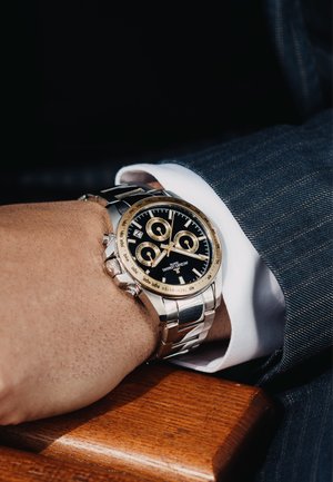 Montre chronographe en acier inoxydable argenté avec un cadran noir, des accents dorés et plusieurs sous-cadrans, portée au poignet avec un costume.