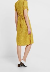 Una donna indossa un vestito giallo a maniche corte, con un motivo, lungo fino al ginocchio, e scarpe con tacco a blocco bianche, mentre si trova di fronte a uno sfondo neutro.