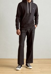 Sudadera con capucha negra y cordones blancos, bolsillo canguro en la parte delantera, combinada con pantalones negros de corte ajustado y zapatillas negras, de pie sobre un suelo de madera.