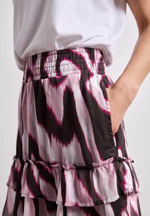 Dłoń kobiety w kieszeni różowo-czarno-białej spódnicy we wzór z falbanką i elastycznym pasem, połączona z białym topem.