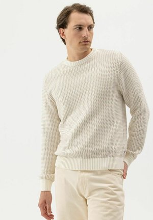 Mann trägt einen cremefarbenen, strukturierten Strickpullover und hellbeige Hosen, steht mit einer Hand in der Tasche und schaut seitlich vor einfachem Hintergrund.