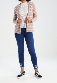 Randig rosa kavaj med sjalkrage, över en vit spetsblus, i kombination med blå smala jeans och svarta sneakers med fransiga fållar.