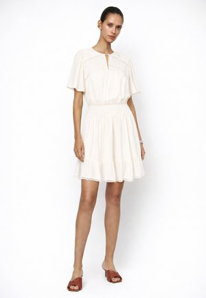 Vrouw draagt een korte mouwen, crèmewitte jurk met een geplooide taille en een geribbelde zoom, gecombineerd met bruine open-toe sandalen met hak.