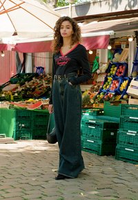 Vrouw met krullend haar draagt een zwarte top en wijde jeans en staat naast kleurrijke fruitkruizen op een buitenmarkt.
