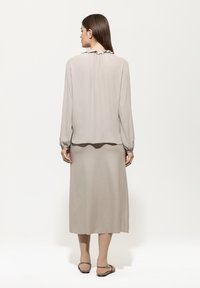 Frau mit dem Rücken zu sehen in einer hellbeigen Bluse mit langen Ärmeln und Rüschenausschnitt, passendem Midi-Rock und flachen Sandalen mit offenen Zehen vor einem weißen Hintergrund.