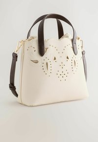 Sac à main couleur crème avec motifs découpés floraux, bord supérieur festonné, poignées brun foncé et bandoulière amovible sur fond neutre.