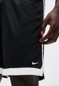 Short de sport noir avec bordure blanche et logo Nike près de l'ourlet, porté par une personne à la peau foncée, main détendue sur le côté.