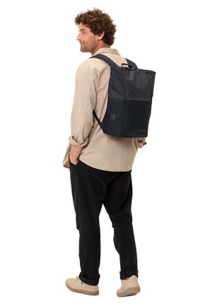 Schwarzer Rucksack mit strukturiertem Finish, rechteckiger Form, Reißverschlussfach und gepolsterten Trägern. Auf einer Person zu sehen, die ein helles Hemd trägt.