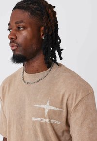 Camiseta marrón de manga corta con un logo blanco en la parte izquierda del pecho, combinada con un collar de cadena plateada y cabello rizado texturizado.
