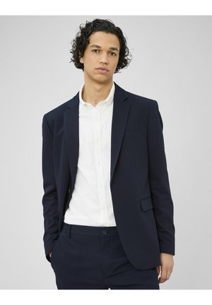 Jeune homme aux cheveux bouclés portant un costume bleu marine et une chemise blanche à boutons, se tenant devant un fond gris clair uni.