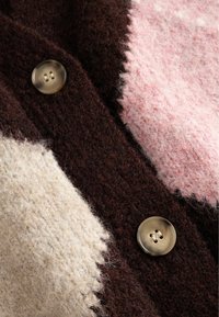Adītais kardigāns ar gaiši brūnām, brūnām un rozā daļām. Izgatavots no teksturētas vilnas materiāla un ar lielu, apaļu, brūnu pogu.