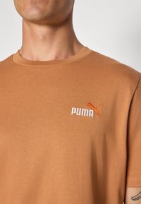 Brun bomull crewneck t-skjorte med en hvit og oransje brodert "PUMA"-logo på venstre bryst. Glatt tekstur og vanlig passform.