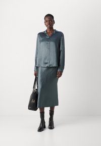 Satin sæt i teal med skjorte og nederdel, der har krave og knaplukning. En sort lædertaske og ankelstøvler fuldender looket. Glat tekstur.