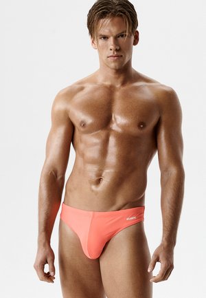 Muskularny mężczyzna z mokrą skórą, ubrany w koralowe kąpielówki z logo "ATLANTIC", stojący na jednolitym jasnym tle.