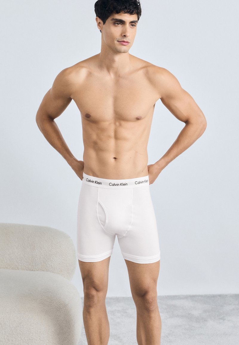 Bijele pamučne bokserice s crnim pojasom "Calvin Klein", pripijenog dizajna i glatke teksture, koje nosi muški model.