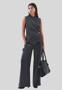 Grå ärmlös omslagstopp med spänne, i kombination med vida grå byxor. Modellen håller en svart handväska med spänne.