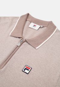 Polo shirt marrone con zip e texture a maglia; presenta un colletto a contrasto e una piccola toppa con logo sul petto.