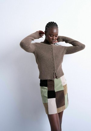 Femme aux cheveux tressés portant un cardigan marron boutonné et une jupe patchwork taille haute dans des tons neutres, posant devant un fond blanc.