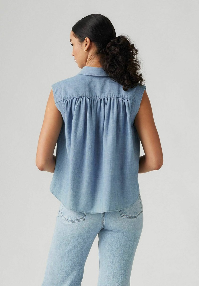 Chemise sans manches bleu clair avec un col, plis froncés au dos et ourlet arrondi. Portée avec un jean bleu clair, le tissu semble doux et aérien.