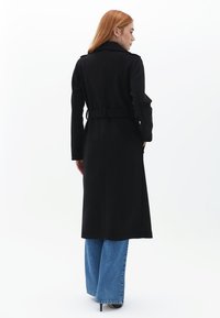 Cappotto lungo nero realizzato in materiale morbido, con cintura removibile, colletto sagomato e spacco posteriore. Abbinato a jeans a gamba larga blu.