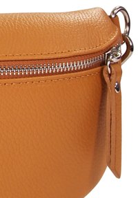 Close-up van de hoek van een tan leren tas, met een zilveren rits, gestikte rand en een aangehechte leren trekstrip met metalen ring.