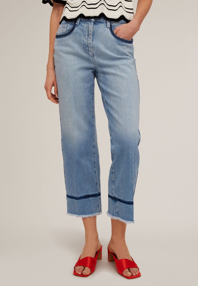 Jeans cortos de color azul claro con dobladillo deshilachado y acentos de color azul oscuro. Llevados con sandalias de tacón abierto rojas. La parte superior tiene un diseño ondulado en blanco y negro.