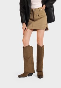 Stivali western in suede marrone con punta a punta e un'ampia canna ondulata. Abbinati a una minigonna khaki e una giacca nera oversize.