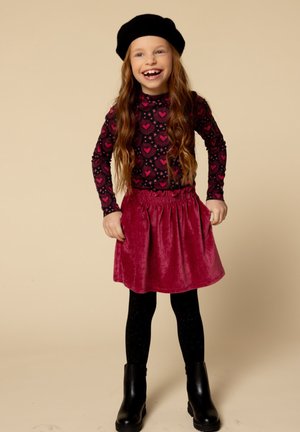 Zwarte lange mouwen top met een levendig roze hartpatroon, gecombineerd met een roze fluwelen rok, zwarte panty’s en zwarte enkellaarsjes.