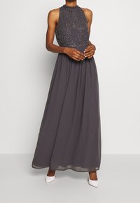 Lång grå aftonklänning med hög hals, utsmyckad bodice med pärlbroderi, och en flödande chiffonskjol som kombineras med vita klackar.