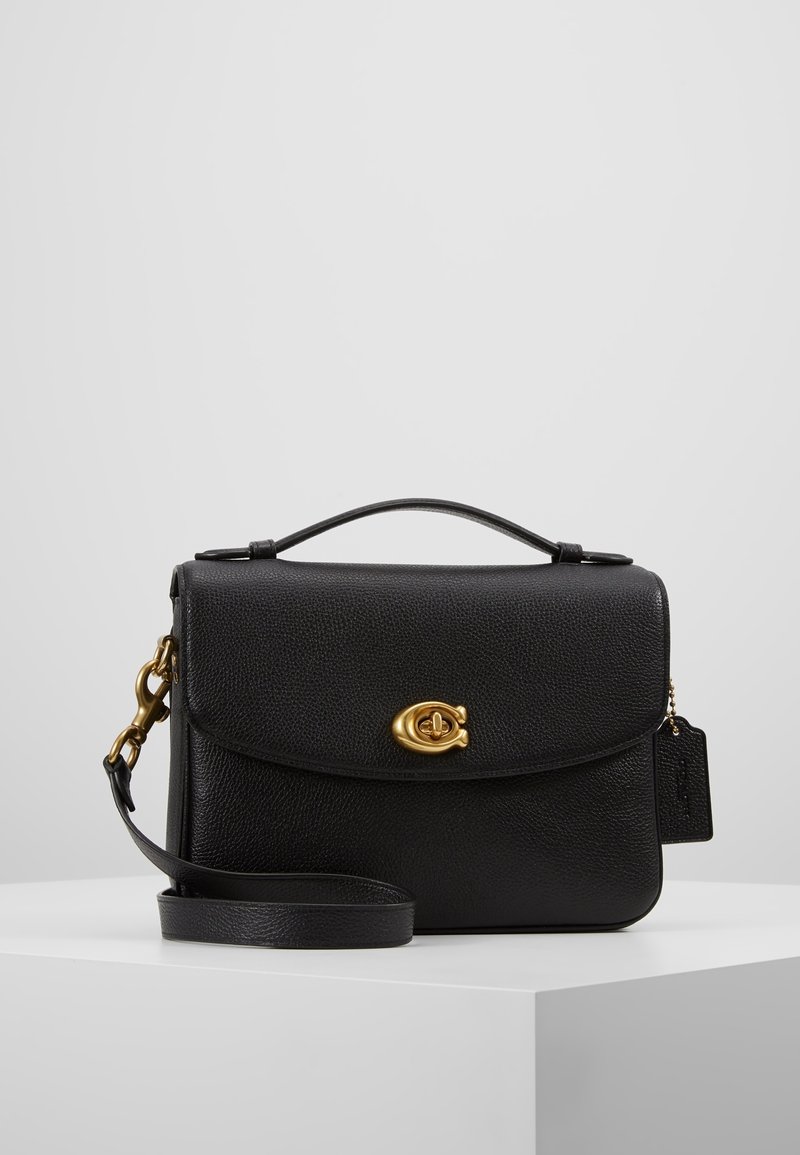 Sac bandoulière en cuir noir avec fermeture à tourniquet dorée, poignée supérieure, sangle réglable et étiquette en cuir suspendue sur une surface blanche.