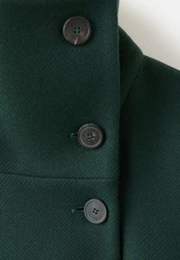 Cuello y parte delantera de abrigo de lana verde oscuro con tres grandes botones negros cosidos con hilo verde a juego.