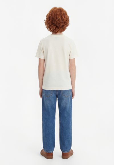 Enfant aux cheveux roux bouclés, debout de dos, portant un t-shirt blanc uni à manches courtes, un jean bleu et des chaussures marron sur fond blanc.