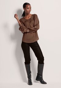 Braune Bluse mit langen Ärmeln und gerefftem Ausschnitt, kombiniert mit schwarzen, eng anliegenden Hosen und hohen schwarzen Lederstiefeln mit Reißverschlüssen.