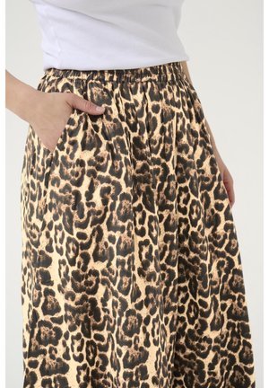 Mano de mujer en el bolsillo de una falda de estampado de leopardo con cintura elástica, combinada con una blusa blanca sin mangas sobre un fondo claro.
