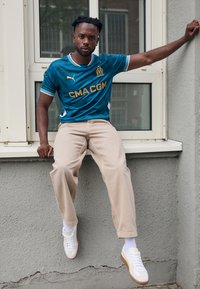 Maillot de football bleu et blanc avec des accents dorés, pantalon beige, baskets blanches, assis contre un mur gris près d'une fenêtre.