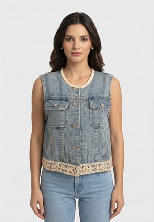 Femme portant un gilet en denim bleu sans manches avec des boutons à l'avant, des poches à rabat et une bordure en dentelle beige à l'ourlet, associé à un jean bleu clair.