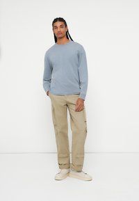 Šviesiai mėlynas ilgas rankovėmis marškinis, lengvi khaki cargo kelnės su šoninėmis kišenėmis ir smėlio spalvos sportbačiai. Modelis stovi prieš paprastą baltą foną.