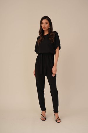 Salopetă neagră cu o siluetă lejeră, mâneci largi și talie cu curea. Poartă sandale negre cu toc, iar materialul are o textură fină.