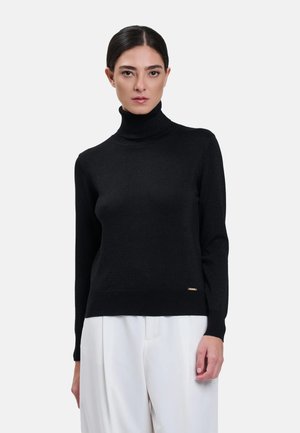 Donna con capelli scuri indossa un maglione nero a collo alto e pantaloni bianchi, in piedi davanti a uno sfondo chiaro semplice, rivolta in avanti.