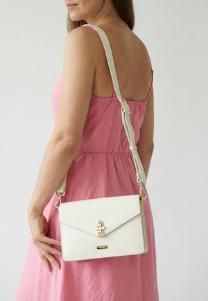 Frau in pinkem Kleid trägt eine weiße Leder-Umhängetasche mit goldenem Verschluss und verstellbarem Trageriemen über der Schulter.