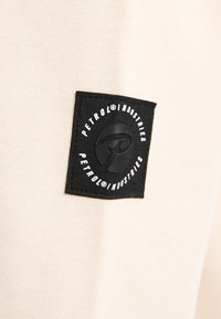 Tela beige con una superficie texturizada que presenta una etiqueta negra cuadrada. La etiqueta muestra "PETROL" en texto blanco y un logotipo circular.