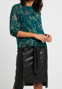 Blouse florale teal avec des manches bouffantes, associée à une jupe en cuir noire taille haute avec des boutons et une taille nouée.