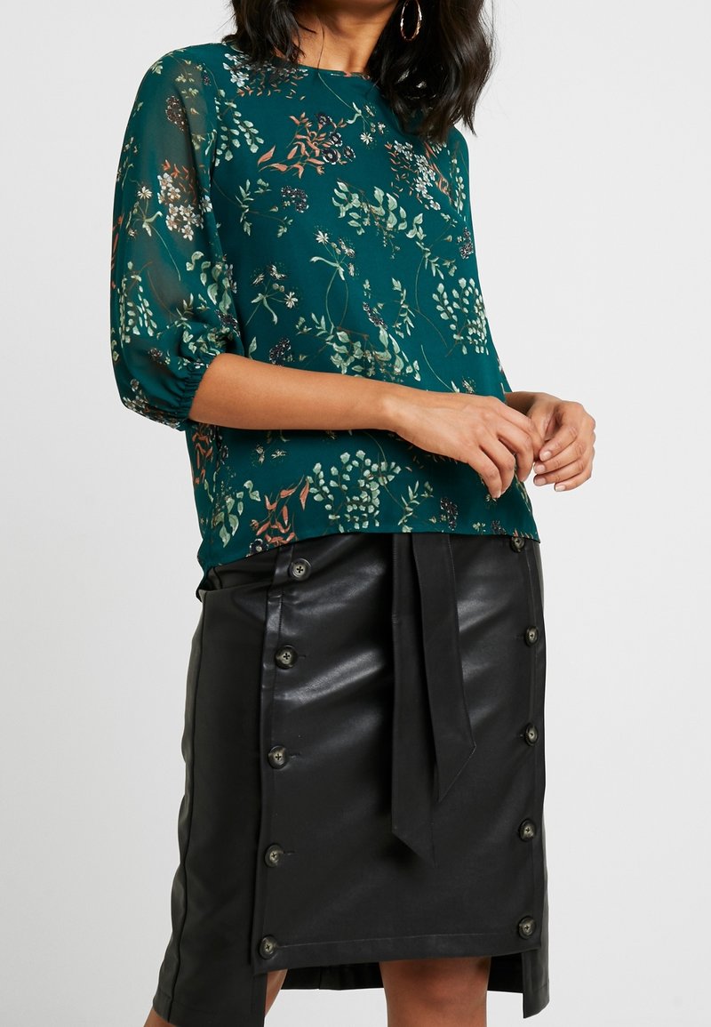 Blouse florale teal avec des manches bouffantes, associée à une jupe en cuir noire taille haute avec des boutons et une taille nouée.