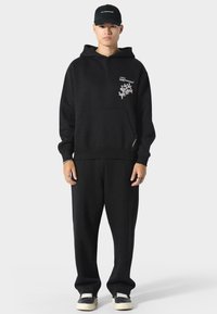 Zwarte hooded sweater met een voorzak, met een witte grafische afbeelding op de linkerborst. Gecombineerd met zwarte, losse broek en sneakers.