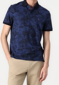 Granatowa koszulka polo z czarnym tropikalnym wzorem, z czarnym kołnierzykiem i krótkimi rękawami, wykonana z lekkiego materiału.