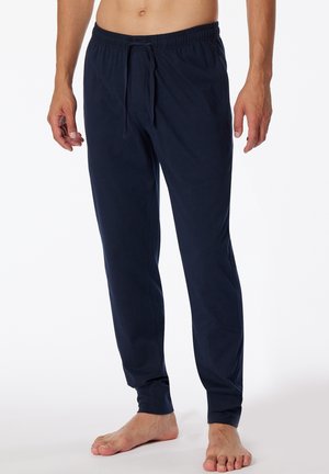 Oben ohne stehende Person in dunkelblauen, elastischen Lounge-Hosen mit Tunnelzug, barfuß vor weißem Hintergrund.