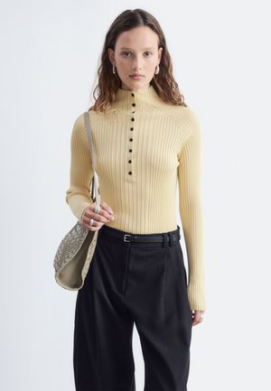 Giovane donna che indossa un top beige a coste a maniche lunghe con bottoni neri, pantaloni neri, anelli d'argento, orecchini a cerchio e porta una borsa a tracolla beige intrecciata.
